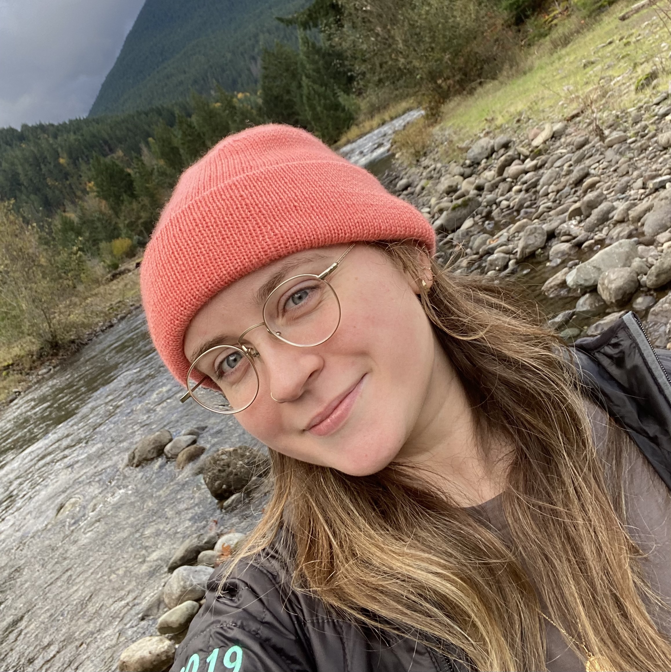 Christina smiling at the camera outside with a river and hills in the background. She is wearing glasses, a pink beanie, and a black jacket.